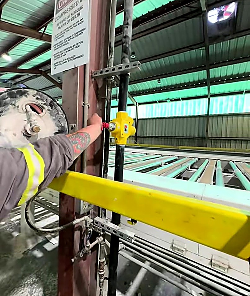 Excel Services tech working a valve on plant piping, rows of roller conveyors running down the hall behind